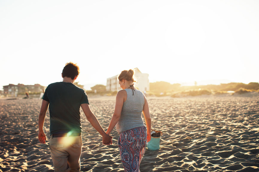 Young Pregnant Woman And Her Husband Enjoying A Sunset On The Beach.